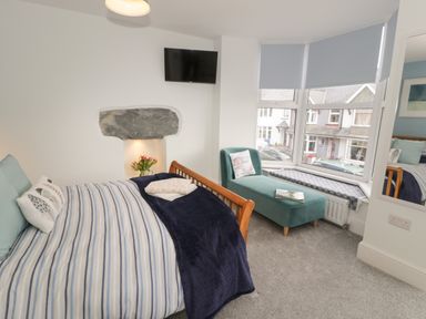 A bedroom with a wooden bed and striped bedding a teal chaise lounge under a bay window and a wall mounted television at Bryn Derfel in Borth-y-Gest near Porthmadog