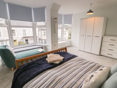 A bedroom with a bed, a green chair, a window seat, white wardrobes, and a chest of drawers at Bryn Derfel in Borth-y-Gest near Porthmadog