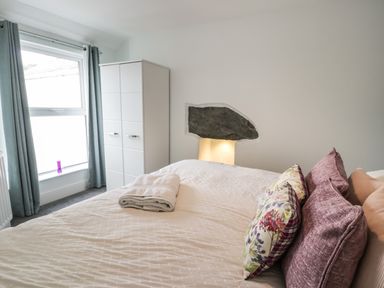 A bedroom with a bed with folded towels on it, pillows, a wardrobe, a window with curtains, and white walls at Bryn Derfel in Borth-y-Gest near Porthmadog