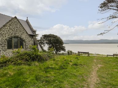 A stone building with a slate roof next to a grassy area overlooking a body of water with benches and hills in the background at Bryn Derfel in Borth-y-Gest near Porthmadog