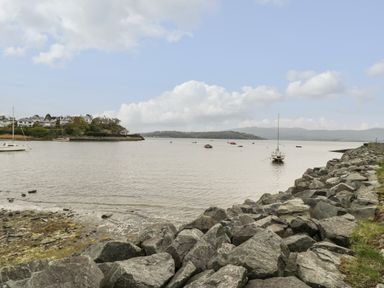 A waterfront with boats anchored near a rocky shore and houses on a hill at Bryn Derfel in Borth-y-Gest near Porthmadog