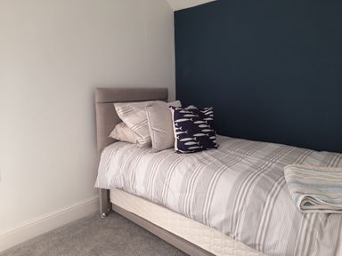 A single bed with striped bedding and multiple pillows against a white and dark blue wall in a bedroom at Bryn Derfel in Borth-y-Gest near Porthmadog