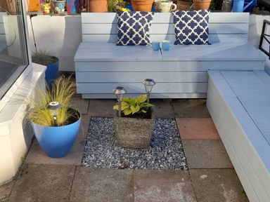 An outdoor seating area with wooden benches painted light blue two patterned cushions two mugs and several potted plants with solar lights