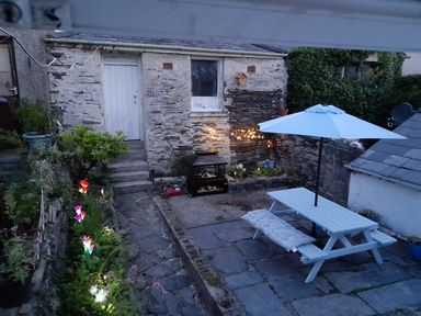 A garden patio with stone paving a blue picnic table and umbrella a chiminea a white door and window on a stone building and illuminated flower lights at Bryn Derfel in Borth-y-Gest near Porthmadog