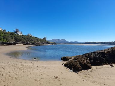 A sandy beach with calm water rocks on the side houses on a hill and mountains in the background at Bryn Derfel in Borth-y-Gest near Porthmadog