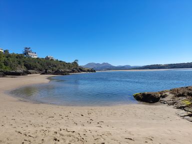 A sandy beach with water and rocky coastline with houses on a hill and mountains in the background at Bryn Derfel in Borth-y-Gest near Porthmadog