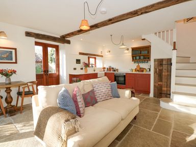 A living room with a beige sofa with cushions a wooden dining table and an open kitchen with red cabinets and shelves at The Little Hay Barn in Bacton