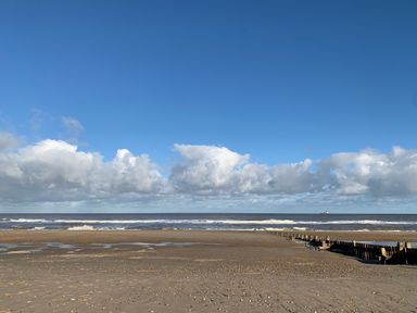 A sandy beach with a wooden breakwater and ocean waves under a blue sky with clouds at The Little Hay Barn in Bacton