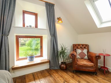 A corner with a leather armchair next to a tall plant and a small red side table by a window with grey curtains at The Little Hay Barn in Bacton