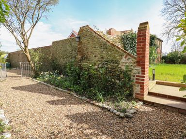 A gravel pathway along a stone and brick wall with plants and flowers next to a grassy area at The Little Hay Barn in Bacton