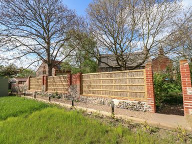 A garden area with grass lawn wooden and brick fencing trees and stone buildings in the background at The Little Hay Barn in Bacton