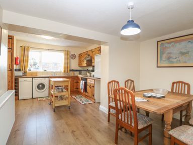 A kitchen with wooden cabinets, a small kitchen island, dining table with chairs, and a window with yellow curtains at Rose Cottage in Downham Market