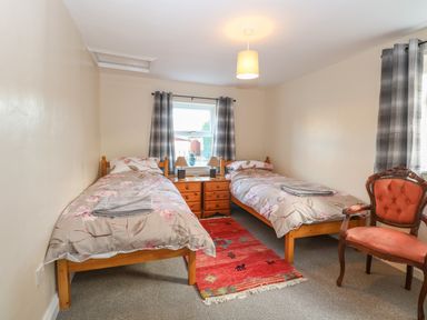 A bedroom with two single beds with floral bedding a wooden bedside cabinet between them a red rug on the floor a window with gray curtains and an upholstered wooden chair at Rose Cottage in Downham Market