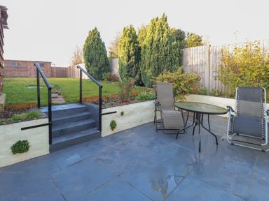 A garden area with stairs leading to grass and trees two reclining chairs and a round glass table on a tiled patio at Rose Cottage in Downham Market