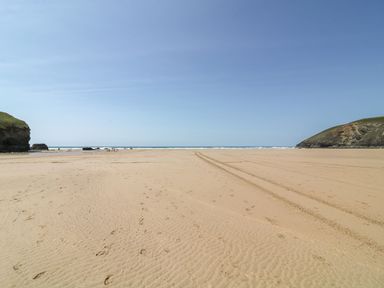 A wide sandy beach with footprints and tire tracks between two grassy cliffs at Pepperpots in Mawgan Porth