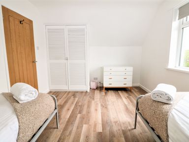 A bedroom with two single beds with rolled towels on them a white chest of drawers a wooden door and a white wardrobe at Pepperpots in Mawgan Porth