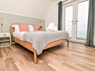 A bedroom with a wooden bed with grey blanket and pink pillows by glass doors with grey curtains at Pepperpots in Mawgan Porth