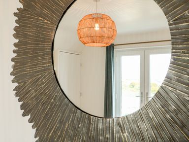 A round mirror with a textured frame reflecting a room with a hanging light fixture and double glass doors at Pepperpots in Mawgan Porth
