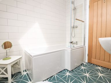 A bathroom with a bathtub shower combination a wooden door a small mirror on a white stool and geometric patterned floor tiles at Pepperpots in Mawgan Porth