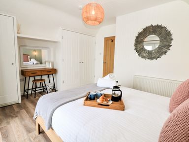A bedroom with a bed with a wooden tray holding coffee cups and croissants and a dressing table with a mirror at Pepperpots in Mawgan Porth