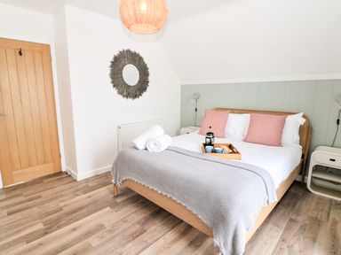 A bedroom with a bed with pink pillows and a gray blanket a wooden door and a circular wall mirror at Pepperpots in Mawgan Porth