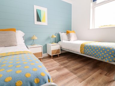 A bedroom with two single beds yellow and blue polka dot blankets nightstands with lamps and abstract art on blue wall at Pepperpots in Mawgan Porth