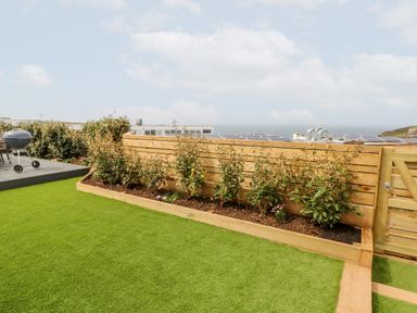 A garden with artificial grass a raised flower bed with plants a wooden fence and a barbecue grill at Pepperpots in Mawgan Porth