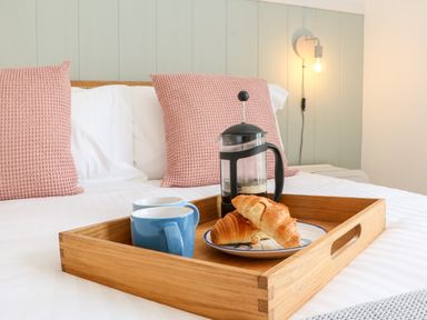 A wooden tray with coffee cups croissants and a coffee press on a bed with pillows and a wall light at Pepperpots in Mawgan Porth