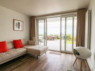 A living room with a beige sectional sofa with red cushions and a sliding glass door opening to a patio at Pepperpots in Mawgan Porth
