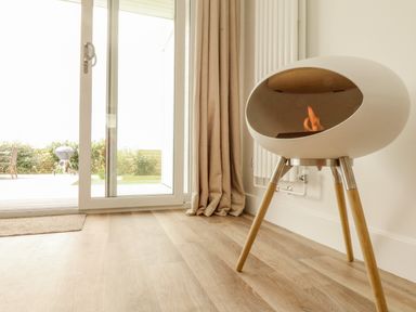 A room with a modern fireplace on wooden legs near a glass door with beige curtains at Pepperpots Mawgan Porth