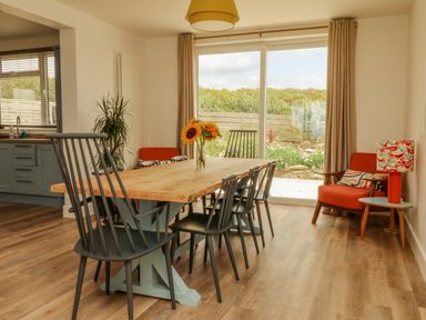 A dining area with a wooden table and black chairs next to a sliding glass door and an orange chair with a side table and lamp at Pepperpots in Mawgan Porth