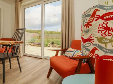 A dining area with a wooden table black chairs orange armchairs and a colorful lamp near sliding glass doors at Pepperpots in Mawgan Porth