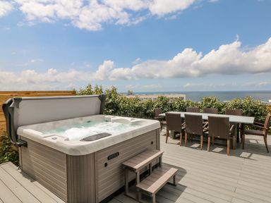 An outdoor deck with a hot tub and a dining table with chairs overlooking the sea at Pepperpots in Mawgan Porth