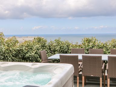An outdoor patio with a hot tub a dining table with wicker chairs and an ocean view at Pepperpots in Mawgan Porth