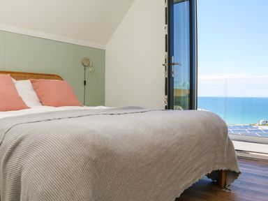 A bedroom with a bed pillows and blanket next to an open door showing a sea view at Pepperpots in Mawgan Porth