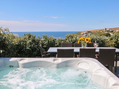 An outdoor seating area with a hot tub a dining table with chairs sunflowers and a bottle of wine with glasses overlooking the sea at Pepperpots in Mawgan Porth