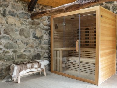 A wooden sauna with glass doors next to a bench covered with a fur throw in a room with stone walls