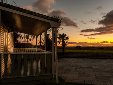 A porch with wooden railing overlooking a paved area and grassy field with a palm tree at sunset at Bennar Fawr in Dyffryn Ardudwy