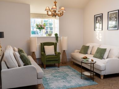 A living room with two beige sofas a green armchair two lamps a chandelier and a window with flowers at Bennar Fawr in Dyffryn Ardudwy