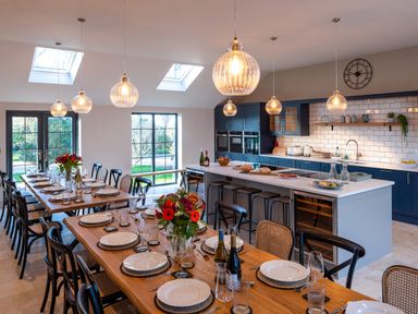 A dining area with two wooden tables set with plates glasses and wine bottles next to a kitchen with blue cabinets white countertops and hanging pendant lights at Bennar Fawr in Dyffryn Ardudwy