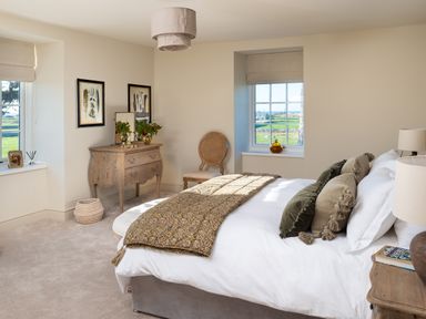 A bedroom with a bed, pillows, a bedside table, a wooden chest of drawers, a chair, and windows showing a green landscape at Bennar Fawr in Dyffryn Ardudwy