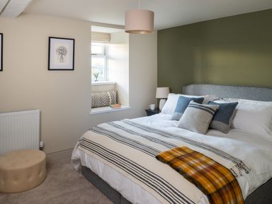 A bedroom with a bed featuring multiple pillows and blankets beside a window with cushions and a small table lamp at Bennar Fawr in Dyffryn Ardudwy