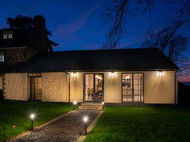 Exterior view of a house with stone and plaster walls lights along a path leading to a door with windows on either side at Bennar Fawr in Dyffryn Ardudwy