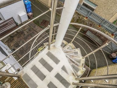 A spiral staircase leading down to a paved courtyard with benches and bins at Awel Y Mor in Criccieth