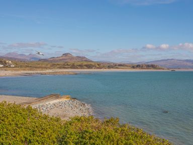 A coastal landscape with a rocky shore a small pier green vegetation and hills in the background at Awel Y Mor in Criccieth