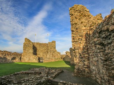 Stone ruins of a castle with green grass and flags on poles at Awel Y Mor in Criccieth