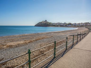 A pebble beach with a railing along a paved walkway and a hill with a castle and houses in the background at Awel Y Mor in Criccieth