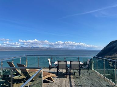A balcony with chairs and a table overlooking the sea and distant mountains at Awel Y Mor in Criccieth