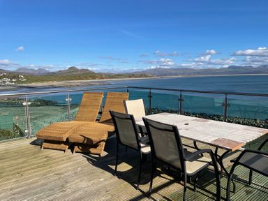 An outdoor deck with chairs a table and loungers overlooking the coast and mountains at Awel Y Mor in Criccieth