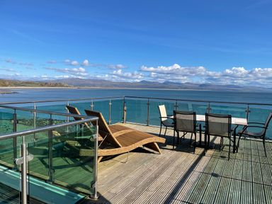 An outdoor deck with lounge chairs and a dining table set overlooking a sea view at Awel Y Mor in Criccieth